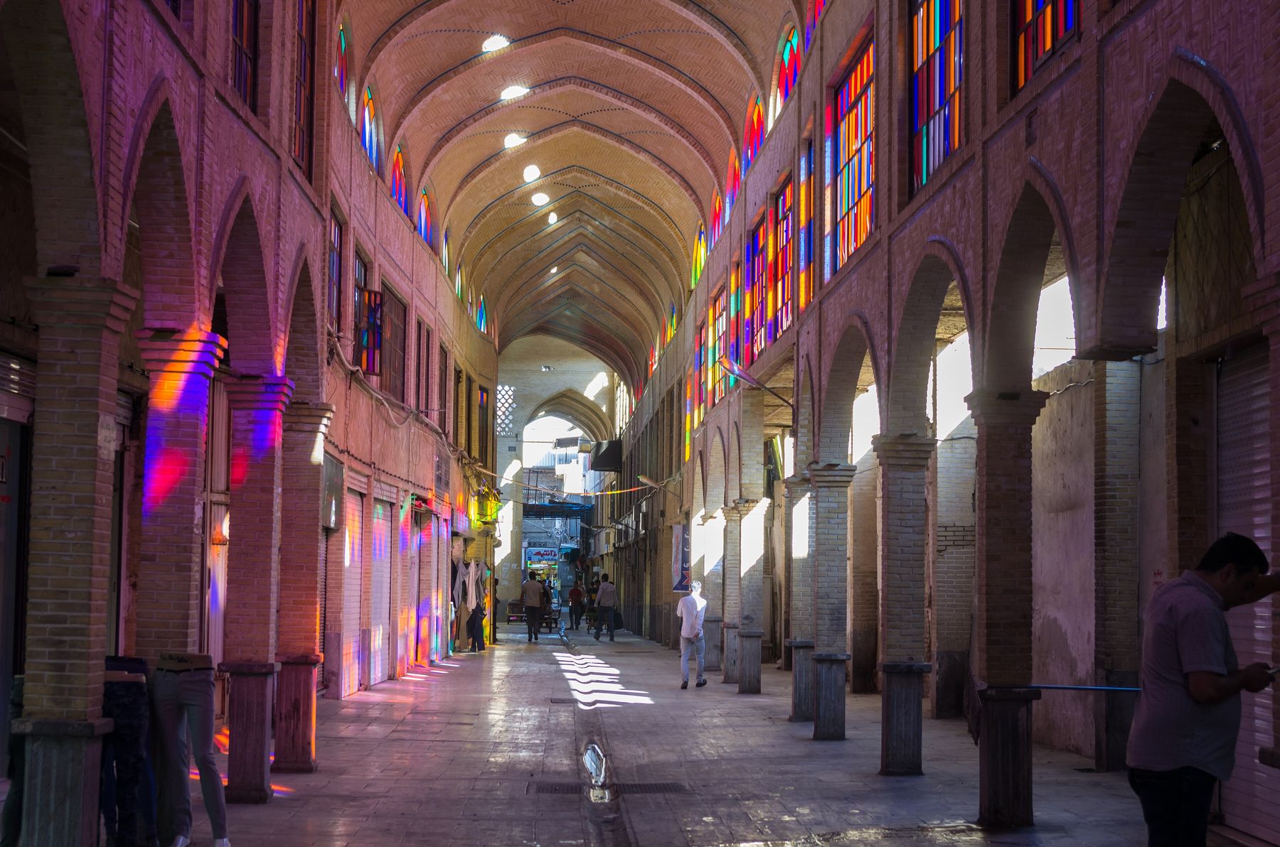 A vaulted, brick-arched corridor in Tehran's Grand Bazaar, lit through stained-glass windows, with most shops shuttered and a few people walking